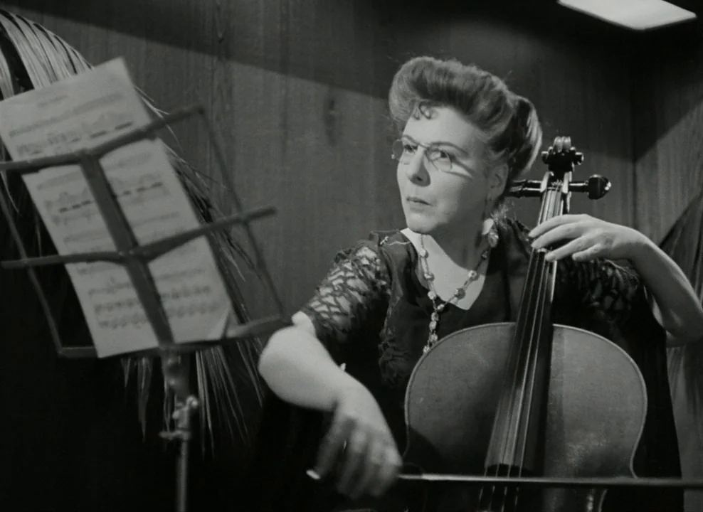 Brief Encounter (1945) movie still: medium — An interior space with wood-paneled walls features a woman playing a cello. She holds a bow to the…
