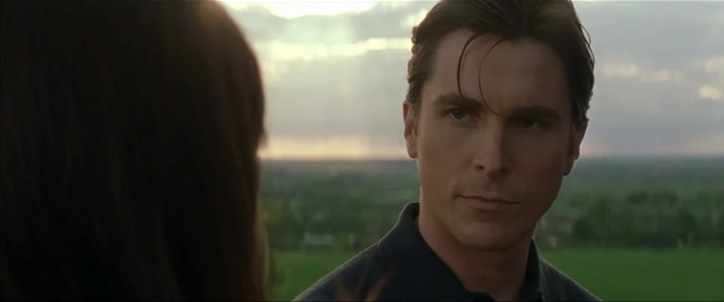 Batman Begins (2005) movie still: closeup — An outdoor grassy field extends to the horizon under a cloudy sunset sky. A man stands in the foreground…