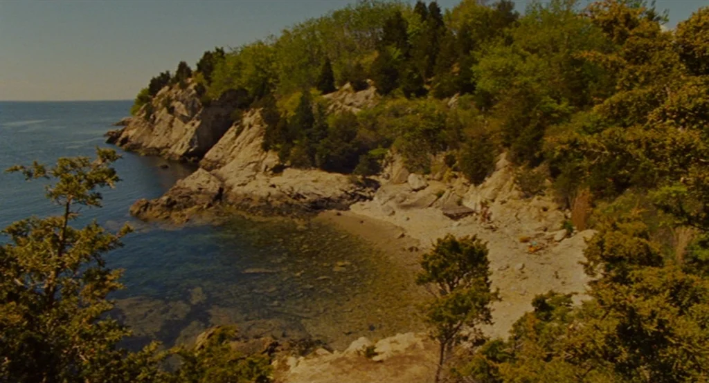 Moonrise Kingdom (2012) movie still: establishing — A secluded rocky cove with a small pebble beach sits beside calm blue water. Trees cover the…