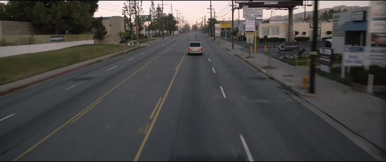 Lowlife (2017) movie still: wide — An empty urban road stretches forward flanked by commercial buildings and utility poles. A single silver minivan…