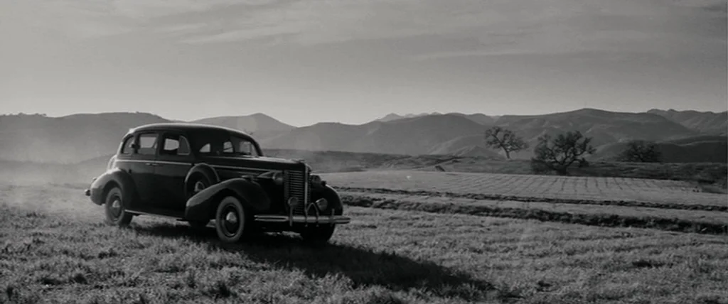 Pineapple Express (2008) movie still: wide — A dark, vintage 1940s sedan is parked in a vast, grassy field. Hilly landscapes and sparse trees are…