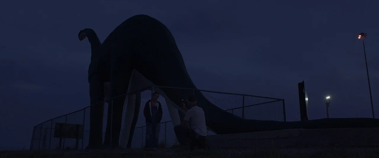 Nomadland (2020) movie still: wide — A giant concrete dinosaur statue stands behind a chain-link fence at night. A man crouches to photograph a…