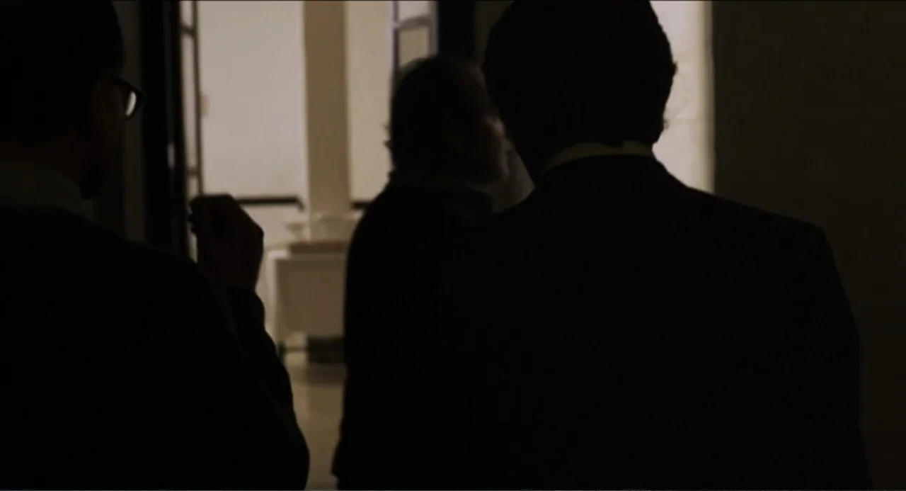 The Holy Girl (2004) movie still: medium — A dimly lit indoor hallway with a bright open doorway in the distance. Three men are standing in the…