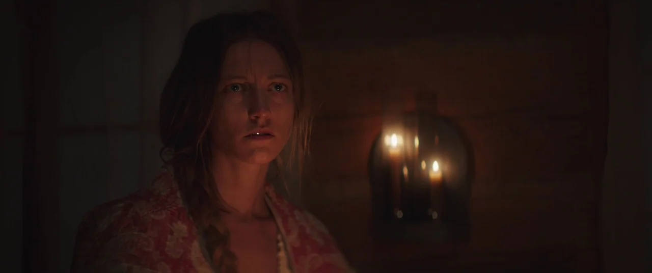 The Wind (2018) movie still: medium — A dimly lit log cabin interior featuring dark wood walls and a flickering wall candle. A young woman looks…