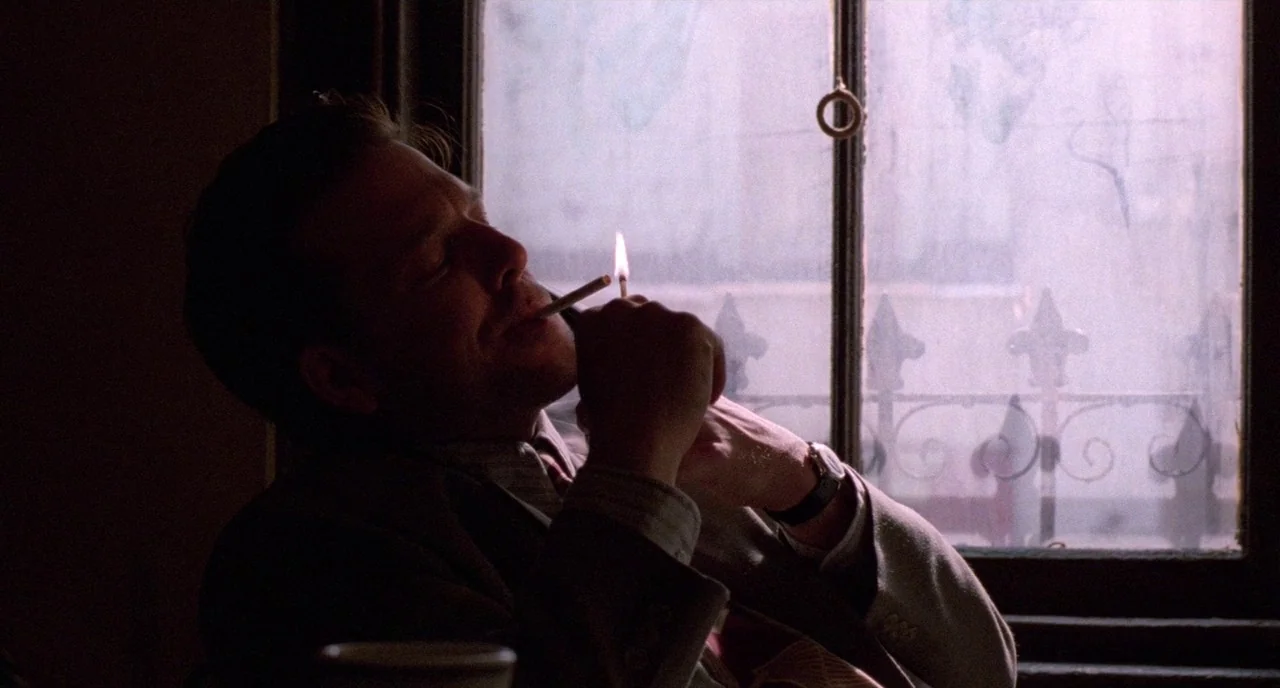 Angel Heart (1987) movie still: medium — A man sits in a diner booth with a window revealing an outdoor metal fence. He tilts his head slightly…