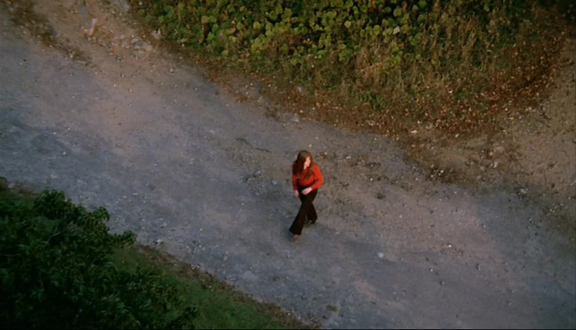 Let’s Scare Jessica To Death (1971) movie still: wide — A rural dirt path winds through foliage near a slightly raised bank. A young woman in a red…