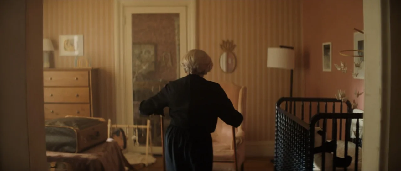 The Front Room (2024) movie still: medium — A quiet nursery features striped wallpaper and a black wooden crib. An older person in dark clothing…