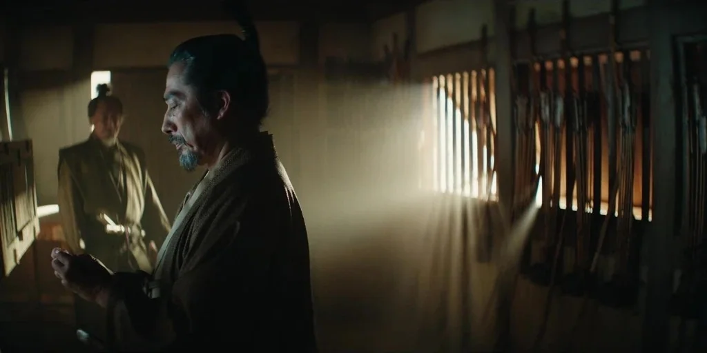 Shogun (2024) movie still: medium — A dimly lit room filled with racks holding wooden bows and arrows. Two men in traditional robes stand within the…