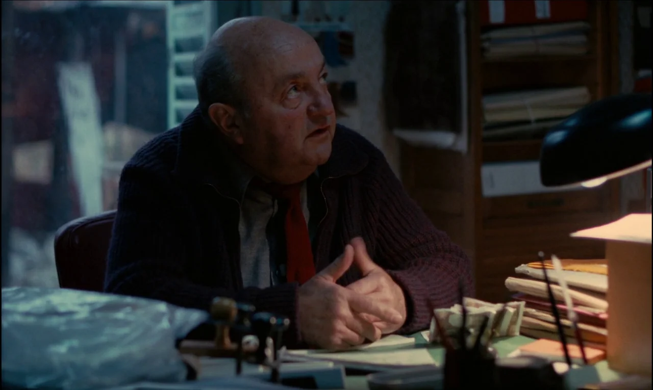 Serie Noire (1979) movie still: medium — A dimly lit workspace cluttered with piles of documents and stacks of paper currency. A man in a sweater…