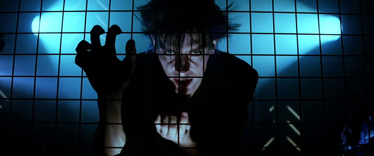 The Hunger (1983) movie still: closeup — A man stands behind a thick metal grid illuminated by harsh blue background light. The man stares directly…