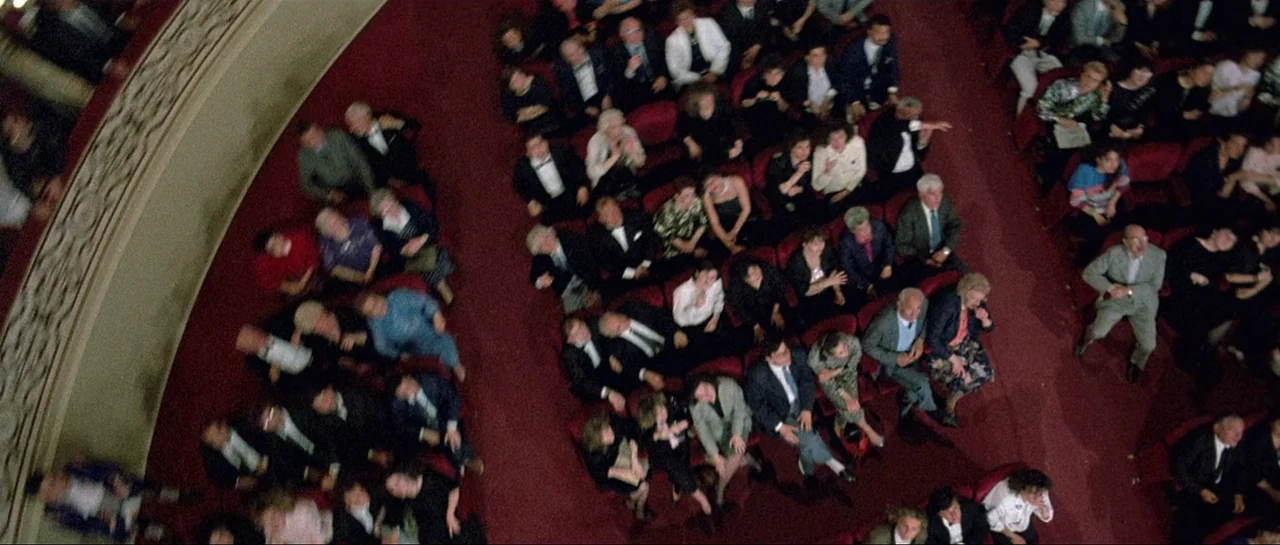 Opera (1987) movie still: establishing — A crowded theater auditorium features rows of red velvet seats filled with spectators. Many people are…