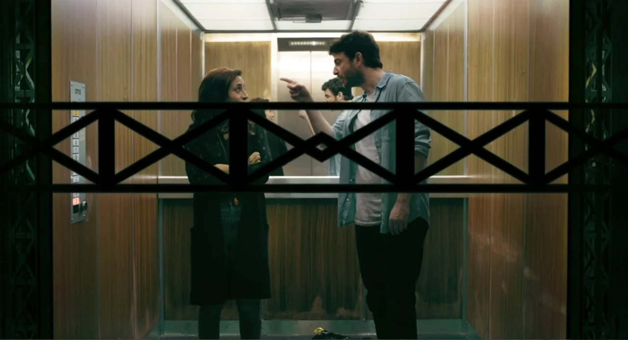 The Lift (2021) movie still: medium — A wood-paneled interior of a modern elevator with metallic buttons on the side wall. A man gestures toward a…
