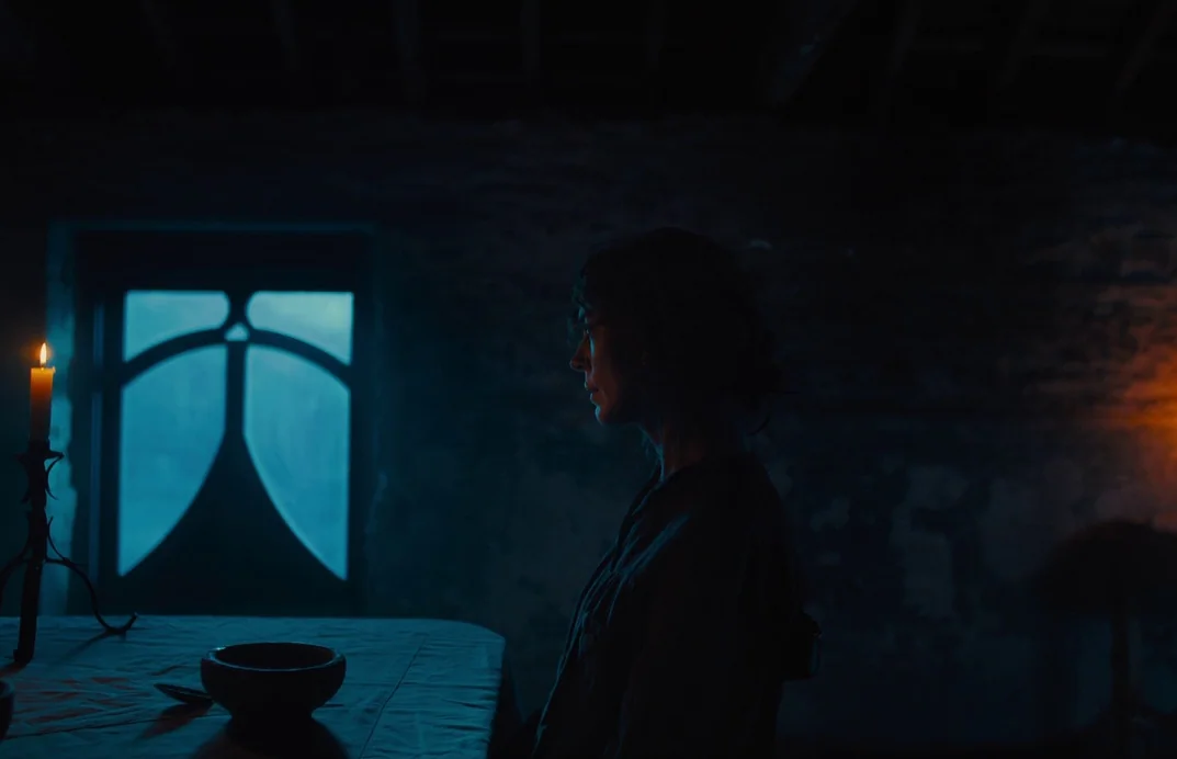 Gretel & Hansel (2020) movie still: medium — A dimly lit room features a wooden table with a bowl and lit candle. A woman sits on a chair in side…