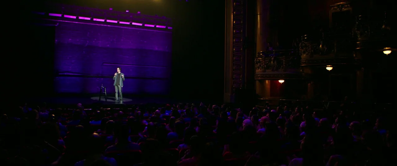 It Chapter Two (2019) movie still: wide — A dimly lit theater auditorium has rows of audience members facing a wooden stage. A lone man stands…