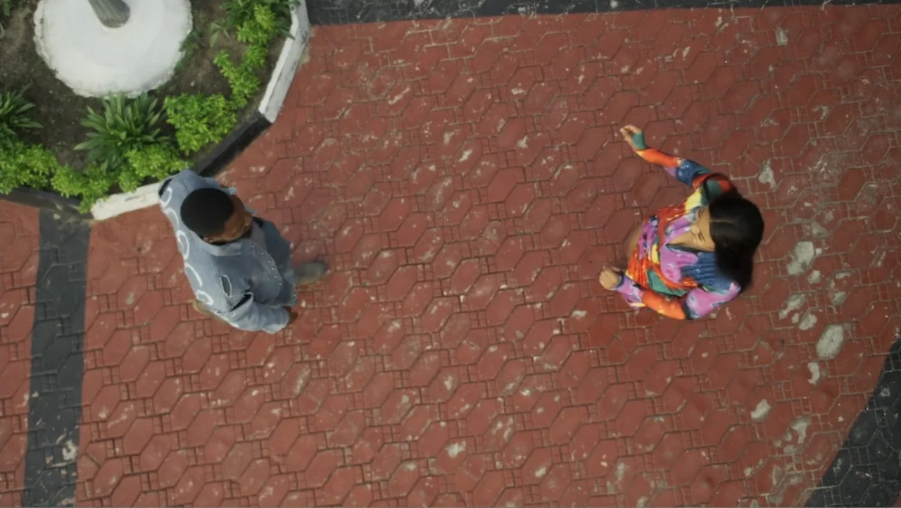 Gangs of Lagos (2023) movie still: wide — An outdoor courtyard features red hexagonal paving stones bordered by white concrete planters. A man and a…