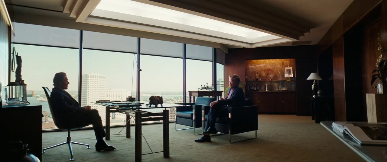 Kinds of Kindness (2024) movie still: wide — A high-rise executive office features panoramic city views and dark wood paneled walls. Two men in…