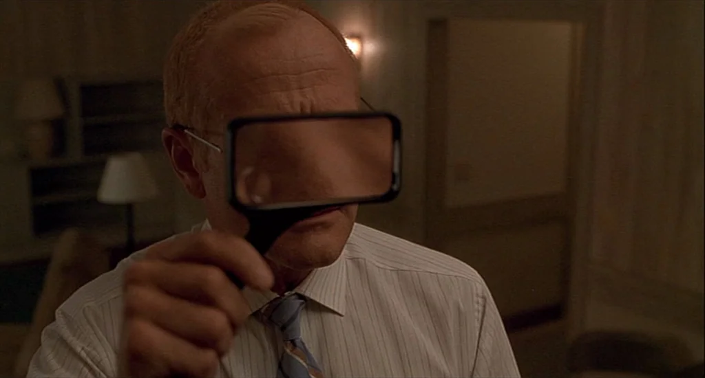 One Hour Photo (2002) movie still: closeup — A dimly lit room with blurred office furniture visible in the background. A man holds a handheld…