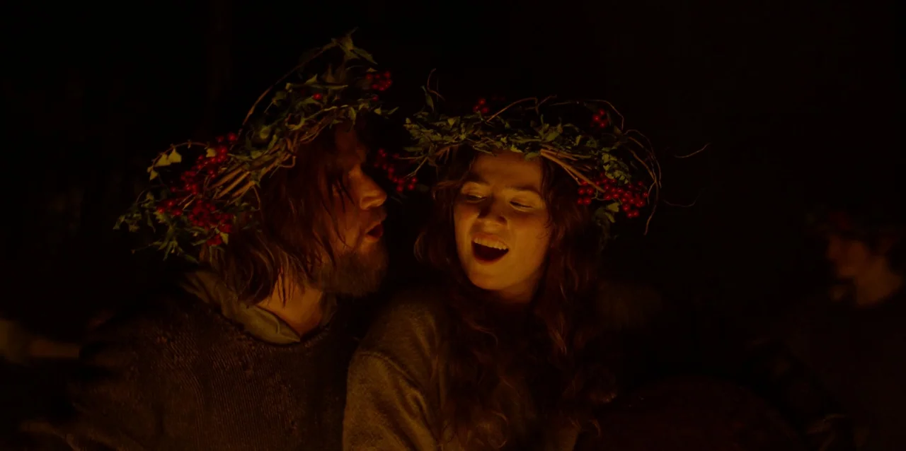 The Northman (2022) movie still: medium — Two people wearing leafy crowns sit close together in a dark exterior space. They are singing while being…