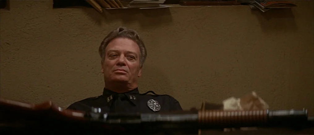 Assault on Precinct 13 (1976) movie still: medium — A dimly lit room with plain walls features a man in a police uniform. He sits behind a desk with…
