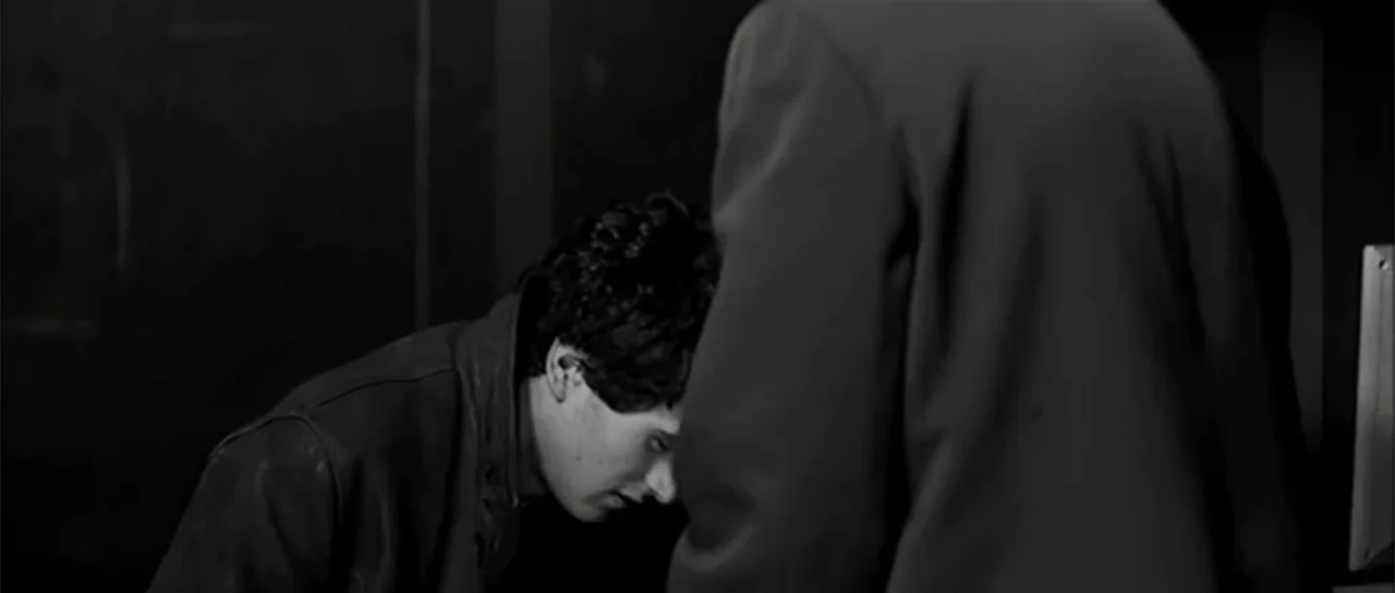 13 Tzameti (2006) movie still: medium — A dimly lit room with high contrast monochrome tones and a tiled wall texture. A young man leans forward…