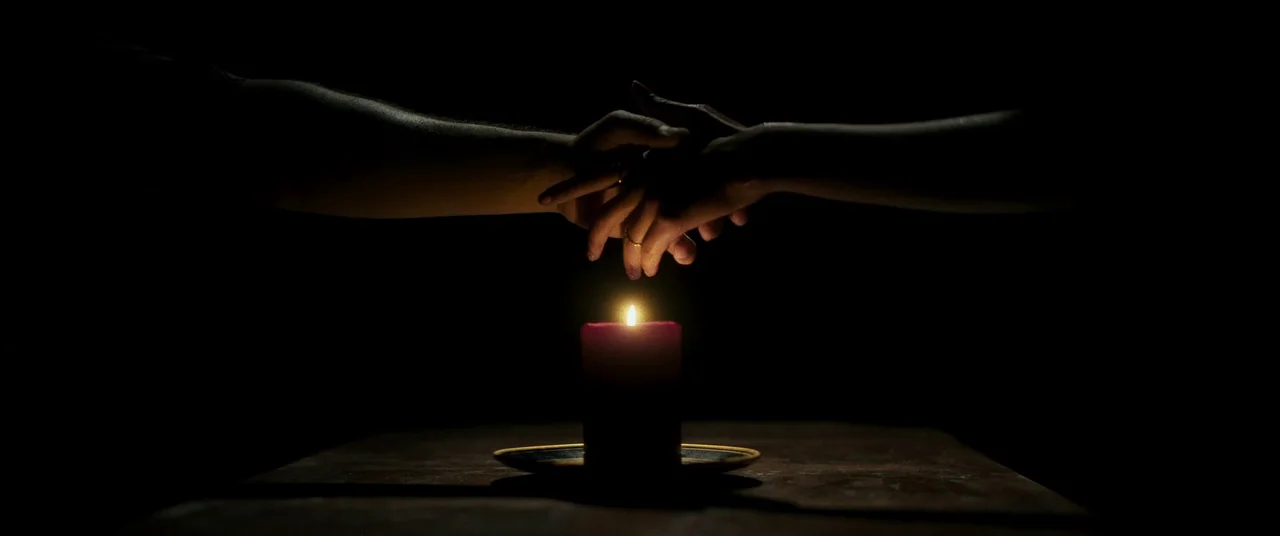 Talk To Me (2022) movie still: closeup — Two human hands reach towards each other in a pitch-black, void-like space. A single lit pillar candle sits…