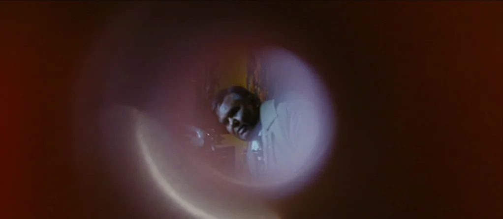 Brick Lane (2007) movie still: medium — A man stands inside a dimly lit space, framed by a circular lens aperture. The subject looks ahead while the…