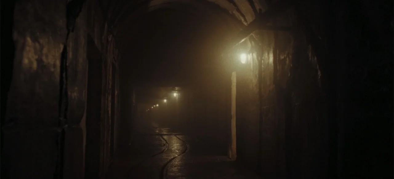 The Tank (2025) movie still: establishing — A dark subterranean tunnel features curved metal tracks embedded in a wet stone floor. Rows of dim amber…