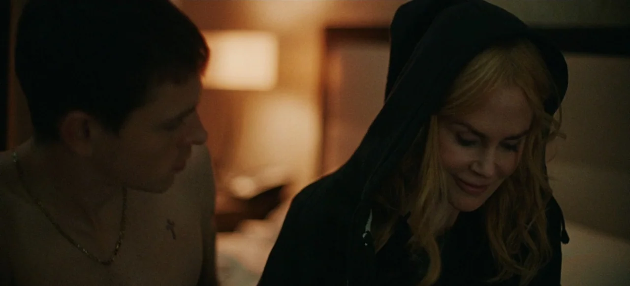 Babygirl (2024) movie still: medium — A dimly lit bedroom features blurred background furniture and a warm lamp glow. A man and a woman stand close…