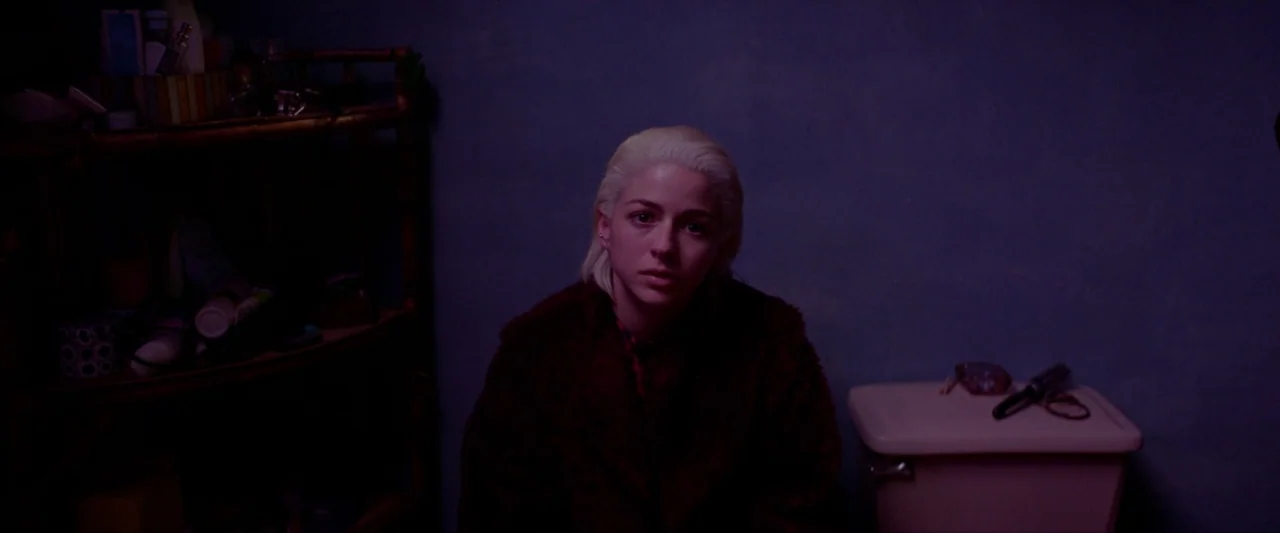 Ema (2019) movie still: medium — A small bathroom with dark violet walls and a wicker shelving unit. A woman sits on the toilet in a dimly lit room.