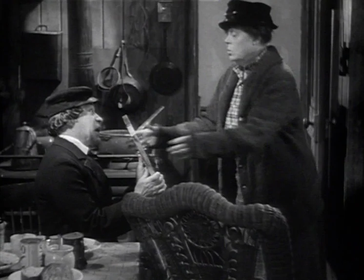 Anna Christie (1930) movie still: medium — A rustic wood-paneled room contains hanging kitchen tools and a table with dishes. One man stands while…