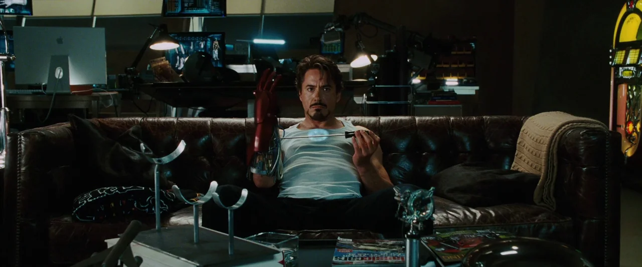 Iron Man (2008) movie still: medium — A man sits on a dark leather tufted sofa in a cluttered industrial workshop area. He holds a screwdriver and…