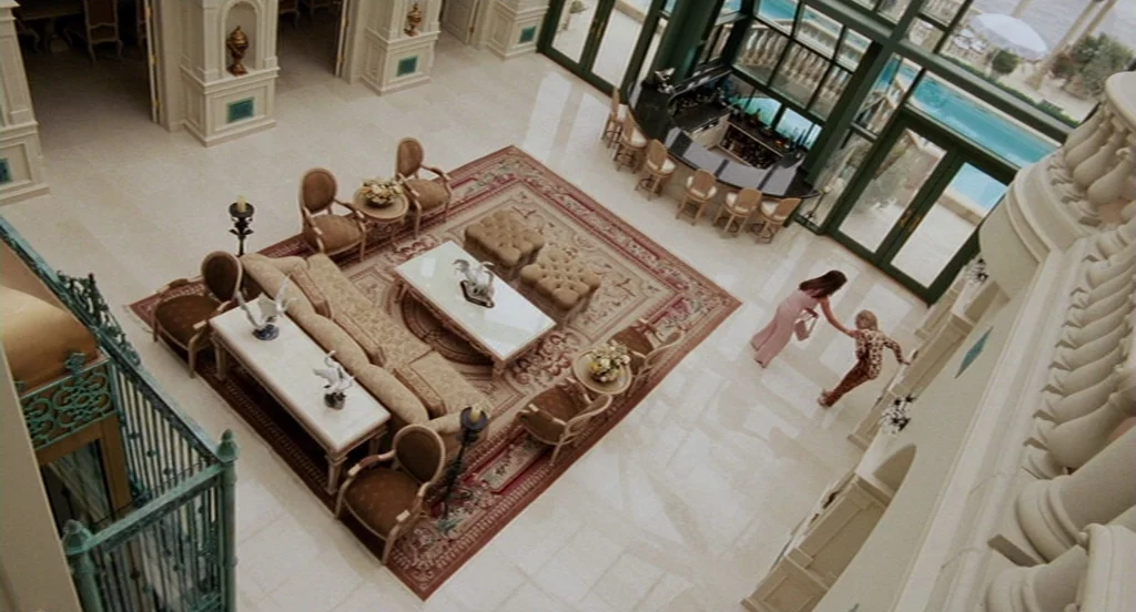 Intolerable Cruelty (2003) movie still: wide — A grand, high-ceilinged room features beige marble flooring and a large, intricate Persian rug. Two…