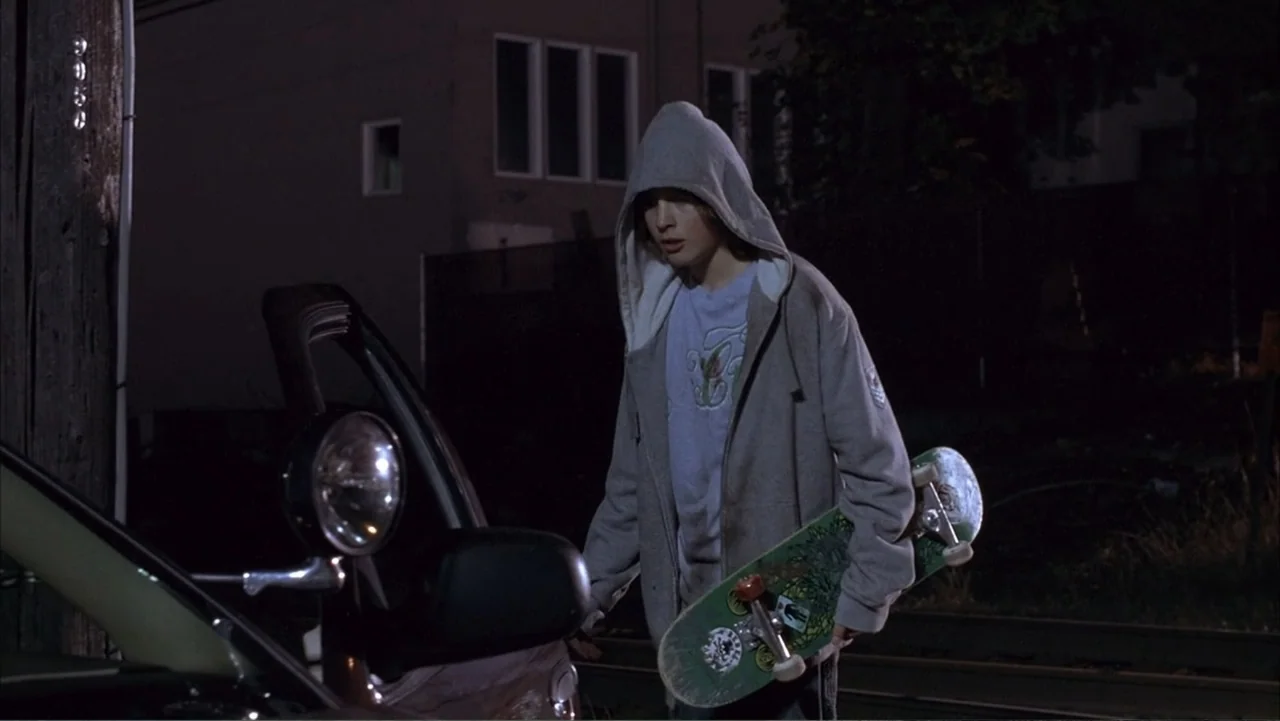 Paranoid Park (2007) movie still: medium — A residential street curb at night next to a dark passenger car. A teenage boy in a hooded sweatshirt…