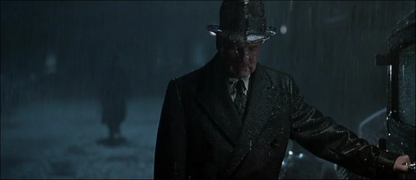 Road To Perdition (2002) movie still: medium — A rain-slicked dark street at night shows a man standing beside a vehicle. The man wears a fedora and…