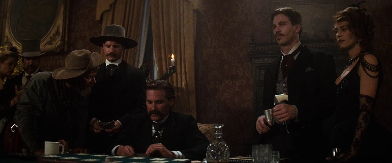 Tombstone (1993) movie still: medium — A dimly lit Victorian-era gambling parlor with patterned wallpaper and ornate wood paneling. Several men and…
