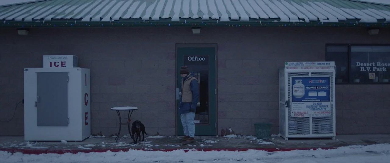 Nomadland (2020) movie still: wide — An RV park office exterior features a brick wall covered in light snow dusting. A man stands at the door with…