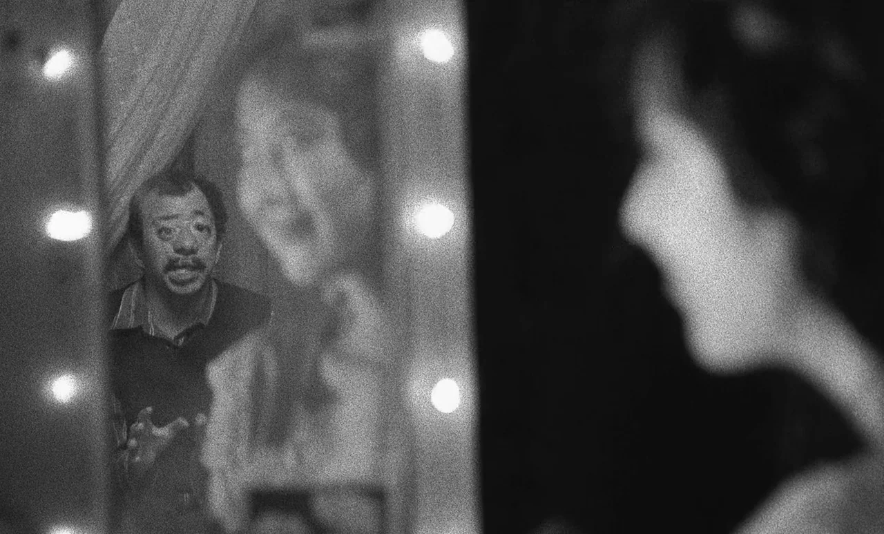 Go Down Death movie still: medium — A man looks into a vanity mirror framed by bright circular light bulbs. A second person is silhouetted in the…