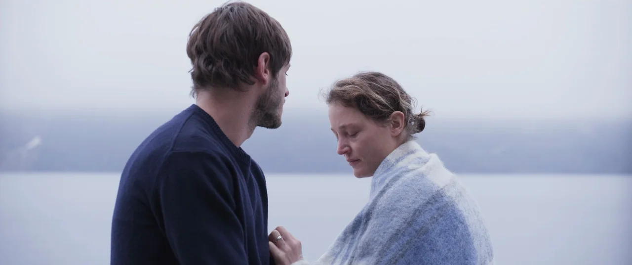 More Than Ever (2022) movie still: medium — A man and woman stand together against the backdrop of a vast, misty lake. The overcast sky blends…