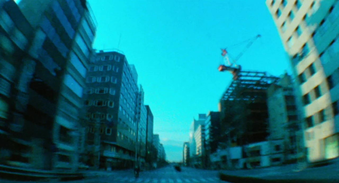 Burst City (1982) movie still: establishing — A wide city street is flanked by tall modern buildings and active construction sites. The perspective…