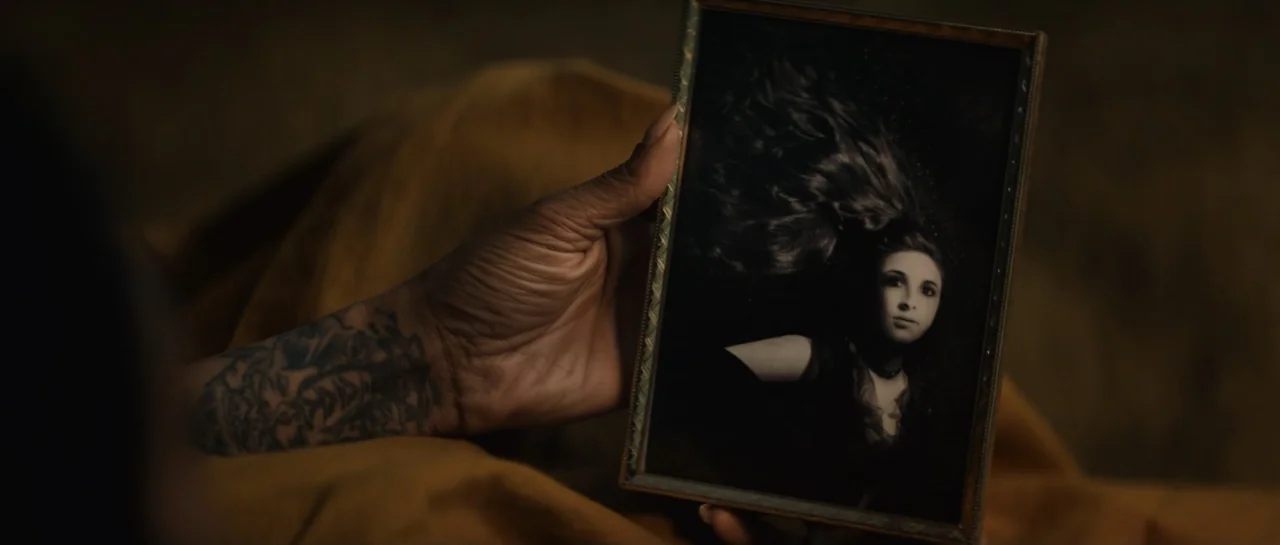 The Front Room (2024) movie still: closeup — A dimly lit interior room features a hand holding a framed monochrome portrait. The photograph depicts…