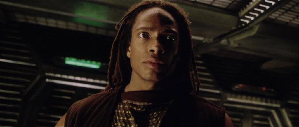 Alien: Resurrection (1997) movie still: closeup — A dimly lit metallic corridor with industrial ribbing on the walls and ceiling. A young man with…