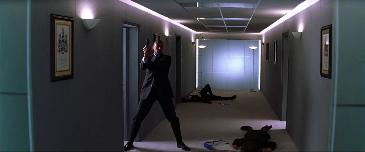 Die Another Day (2002) movie still: fullbody — A modern corporate corridor with gray walls and two bodies lying on the floor. A man in a suit stands…