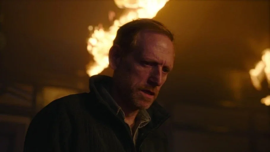 Making of: The Last of Us (2023) movie still: medium — A man stands inside a building filled with smoke and active flames. He looks down while the…
