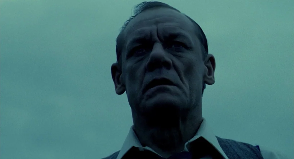 In a Glass Cage (1986) movie still: closeup — A man stands outdoors under a cloudy sky with a uniform blue tint. The man looks forward into the…