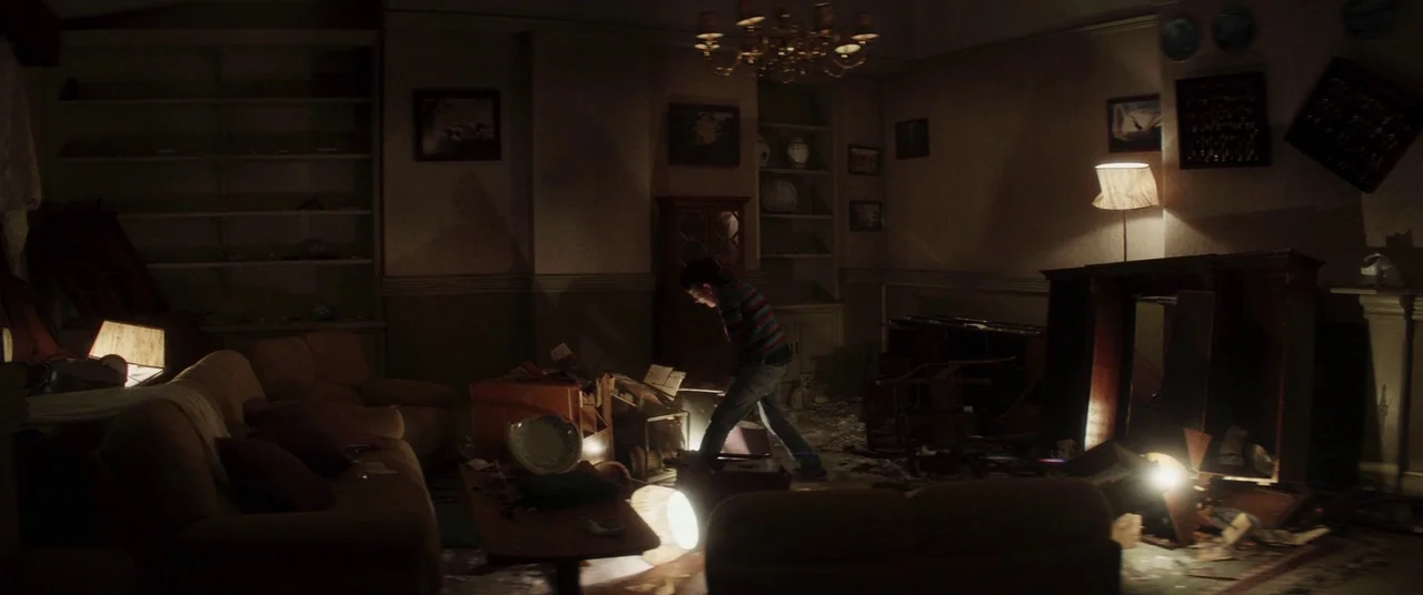 A Monster Calls (2016) movie still: wide — A cluttered, dark living room filled with scattered household items and overturned furniture pieces. A…