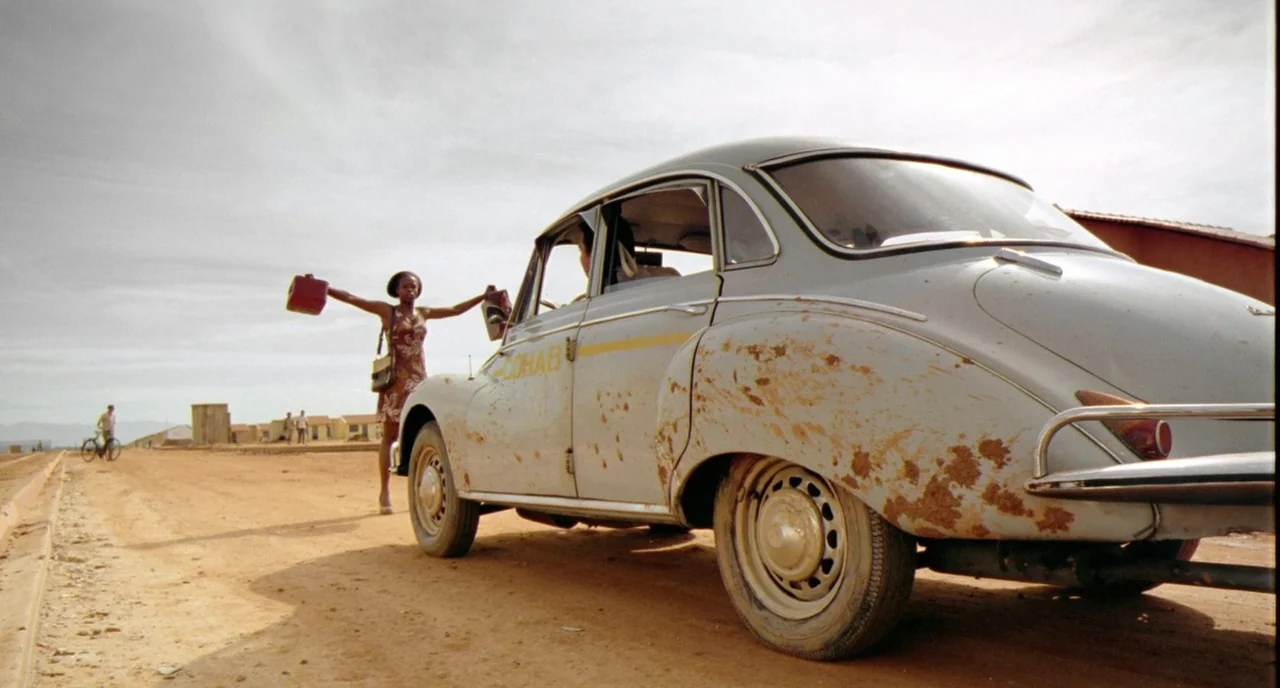 City of God (2002) movie still: fullbody — A dusty unpaved road stretches past small houses under a pale, overcast sky. A woman stands beside a…