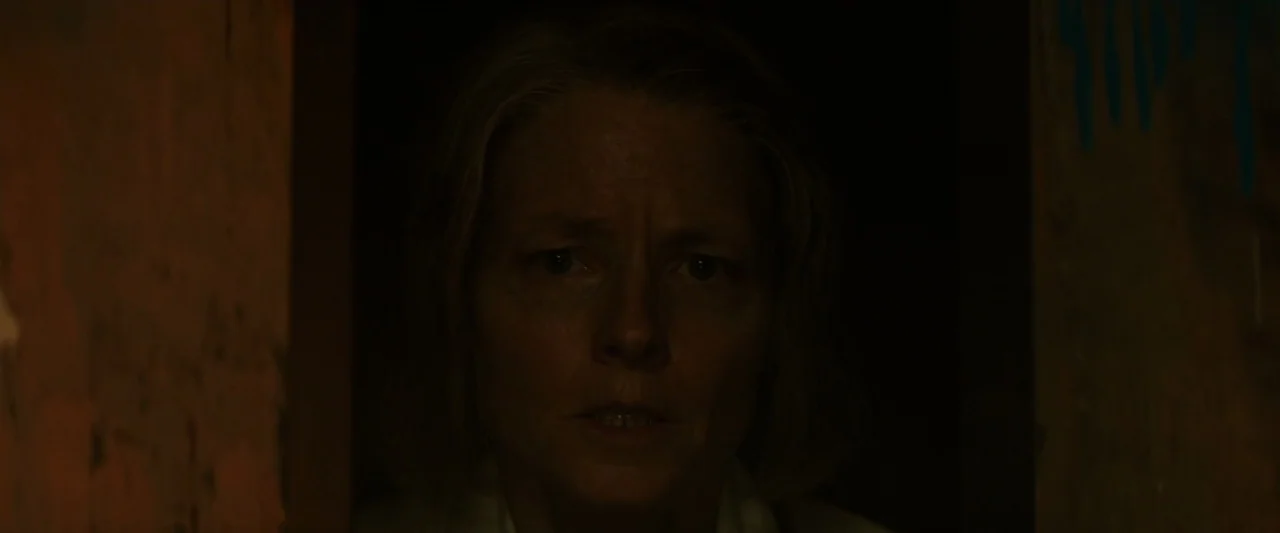 Hotel Artemis (2018) movie still: closeup — A woman stands in a dimly lit, narrow corridor with dark, obscured walls. She gazes ahead from the…