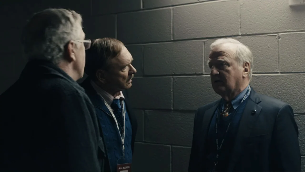 Paterno (2018) movie still: medium — A dimly lit concrete hallway features plain block walls and utilitarian architectural lines. Three middle-aged…