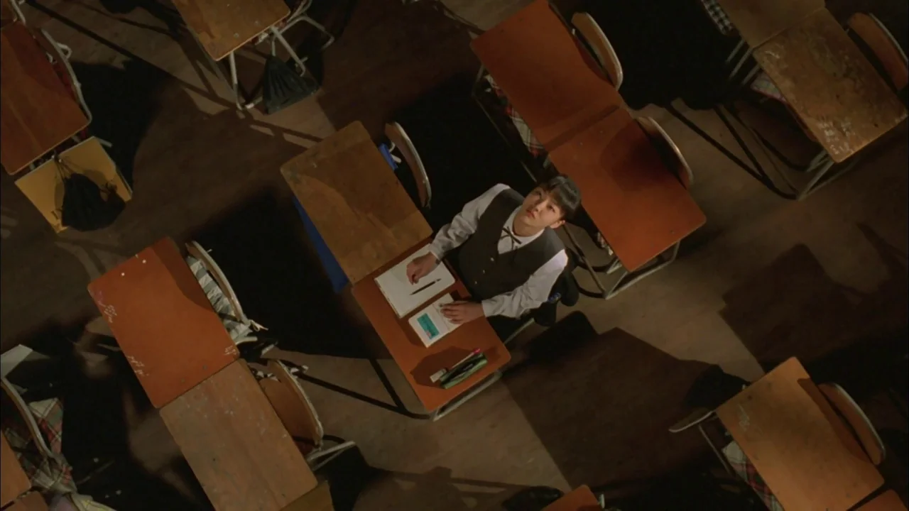 Whispering Corridors (1998) movie still: wide — A rows of wooden school desks are arranged in a dimly lit, empty room. A solitary male student sits…