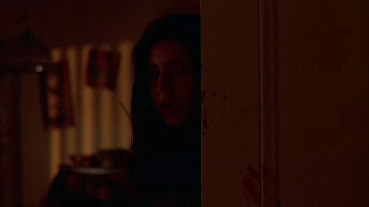 Ginger Snaps (2000) movie still: closeup — A dark indoor space viewed from behind a door frame with visible red stains. A woman with dark hair looks…
